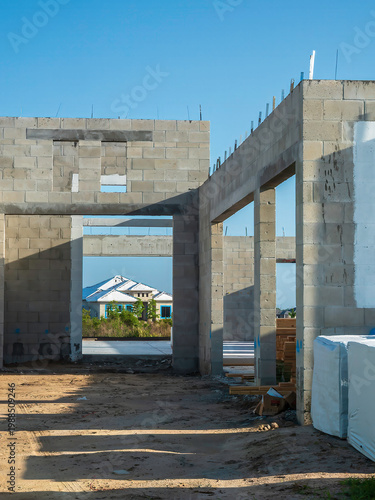 Partial view of the concrete shell of a house under construction, with framed view of a neighboring house in a later stage of construction, in a suburban development on a sunny morning in Florida
