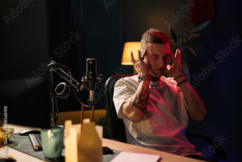 Obraz na płótnie Young adult Caucasian man streaming podcast, touching temples while focusing beside microphone and headphones