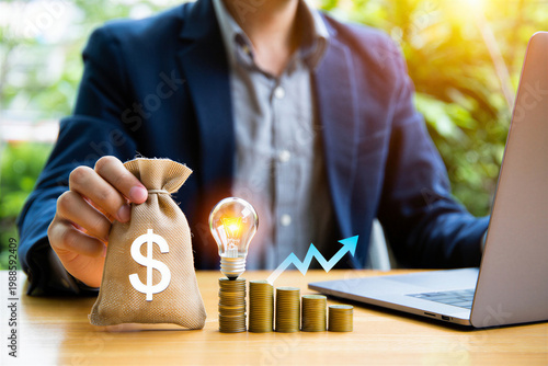 A businessman sitting at a desk with a money bag, light bulb, and laptop, symbolizing a bright financial idea.