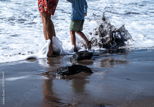 A father and son are walking along the seashore. The waves crash around them as they stroll along the shore. 