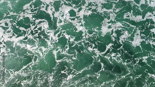 At Sea - Aerial view of foamy waves creating interesting abstract patterns in deep turquoise water. Swirling white foam contrasts emerald ocean texture at open sea.