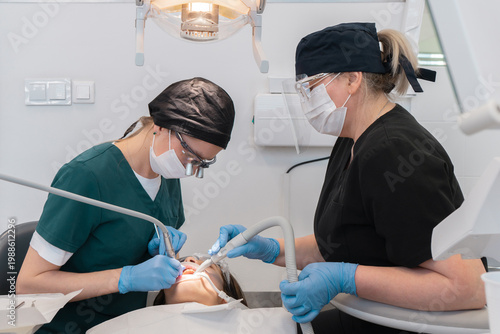 Healthcare specialist team performing dental procedure on patient in medical office. Female dentist and assistant using professional equipment for stomatology treatment and oral health.