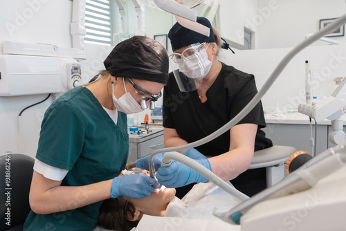 Professional healthcare team performing four-handed dentistry in stomatology clinic. Female dentist and assistant using drill and suction for tooth restoration and oral surgery procedure.