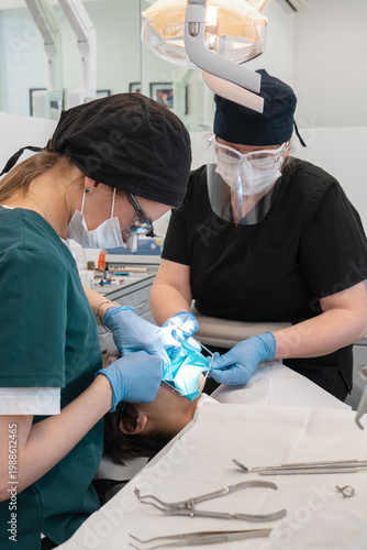 Dentist and assistant installing metal rubber dam frame on patient mouth for tooth treatment. Medical team in protective gear performing professional stomatology procedure in dental clinic.