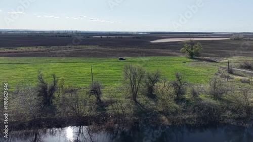High altitude aerial view of blue river reflecting sun near lush green meadow. Dark car parked on field surrounded by agricultural lands and spring trees under clear sky in countryside.
