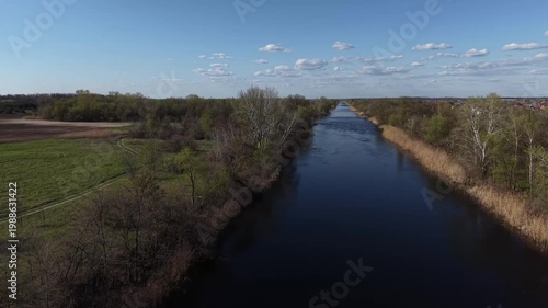 Drone flight along a straight water canal or river. View of blue water, spring trees, coastal reeds and distant village under a clear sky.