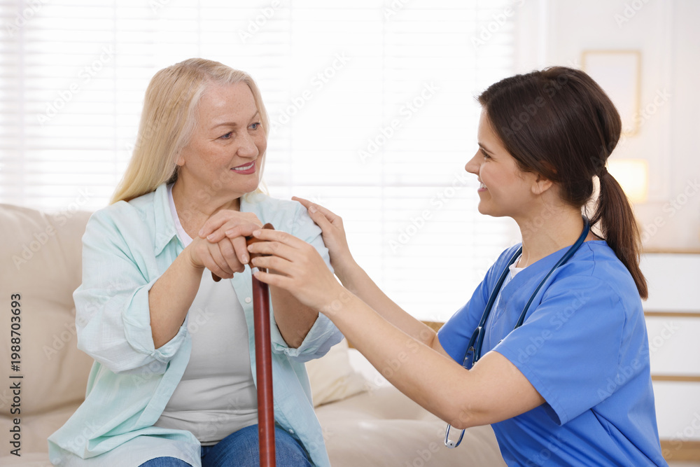 Fototapeta premium Smiling care worker supporting senior woman with walking cane at nursing home