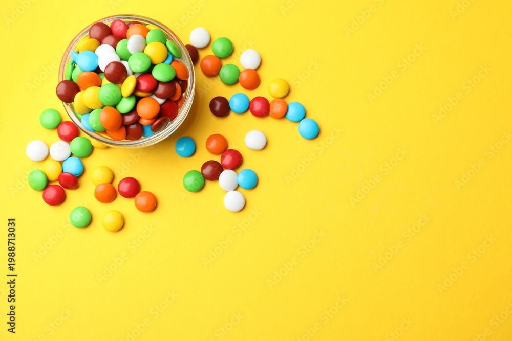 Obraz premium Delicious colorful candies in glass bowl on yellow background, flat lay. Space for text
