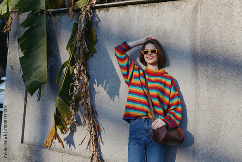 Canvastavla Woman wearing a colorful striped sweater and sunglasses leaning against a concrete wall with tropical leaves, smiling in a casual outfit and jeans in an urban lifestyle travel scene