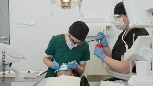 Dentist uses restoration instrument to place composite material into cavity while assistant holds curing light. Professional dental team performing tooth filling procedure in clinic healthcare.