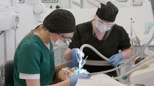 Dentist in loupes and assistant with face shield treating teeth of patient in clinic. Professional medical team using drill and suction during oral surgery or hygiene procedure in hospital.