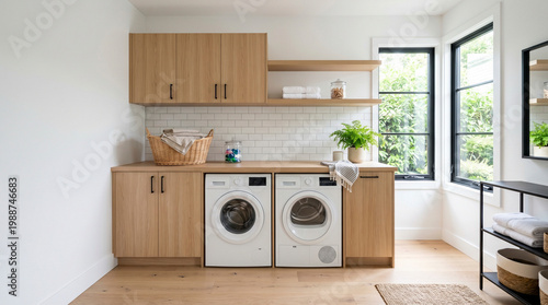 Bright and modern laundry room featuring light oak wood cabinets, white subway tile backsplash, a washing machine, dryer, wicker baskets, large window with black frames.
