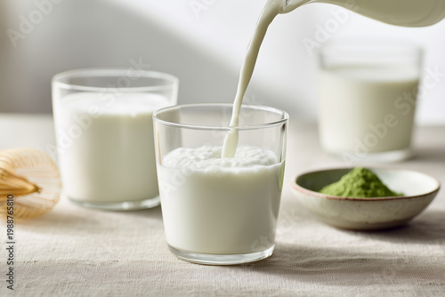 Close up of pouring milk into glass with matcha powder and bamboo whisk on linen background