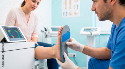 A doctor is examining a patient's foot while discussing custom insoles in a podiatrist office. The room is equipped with medical tools and devices for foot care