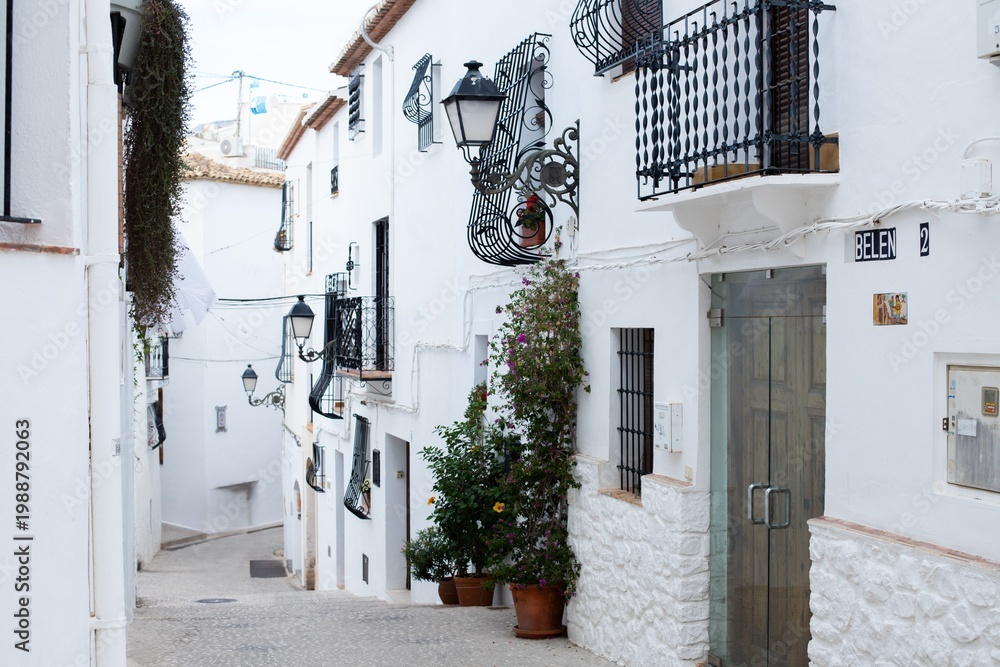 Fototapeta premium Spain, Altea city. Narrow street in a picturesque Spanish village with whitewashed buildings, decorative balconies, and vibrant flower pots lining the pathway