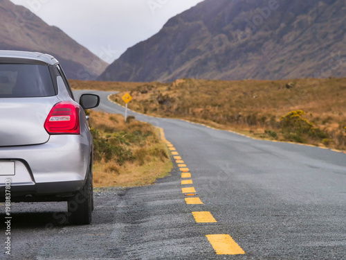 Silver car parked beside rural road. Mountain looms distant under cloudy sky. Road stretches ahead, flanked by dry grass. Connemara area. Ireland travel and tourism.