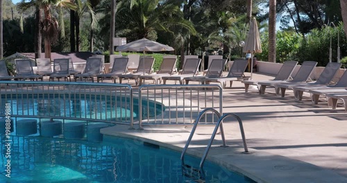 PORTOCOLOM, MALLORCA, SPAIN - OCTOBER, 2025: Empty sun loungers arranged by a swimming pool under midday sun creating sharp contrast and shadows, wide shot