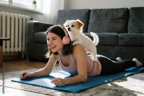 Woman doing yoga at home with a small dog on her back, wearing headphones and smiling