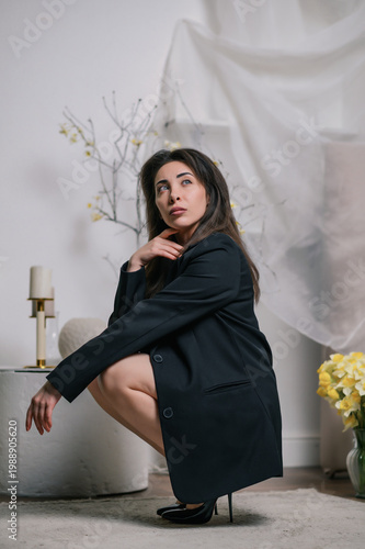 Stylish woman in black blazer posing in minimalist studio interior with soft draped fabric and delicate floral decor, natural light, elegant and thoughtful mood, fashion portrait