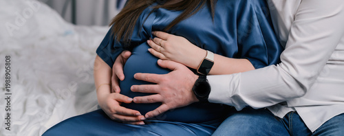 Close-up of pregnant woman in blue dress holding her belly with partner’s hands, soft indoor light, intimate home setting, symbolizing care, love and family expectation for healthcare and lifestyle