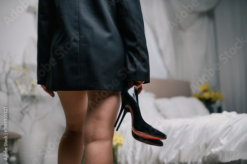 Back view of woman in oversized black blazer holding high heel shoes in minimalist bedroom with soft bedding and neutral tones, stylish fashion lifestyle concept for branding and advertising use