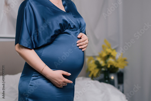 Close-up of pregnant woman in blue satin dress holding her belly, soft indoor lighting, cozy home bedroom background, calm and caring mood, concept of motherhood, prenatal care and family lifestyle