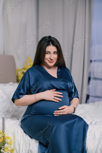 Pregnant woman in blue satin dress sitting on bed, gently holding her belly, cozy home interior with soft light, calm and happy mood, concept of motherhood and family lifestyle