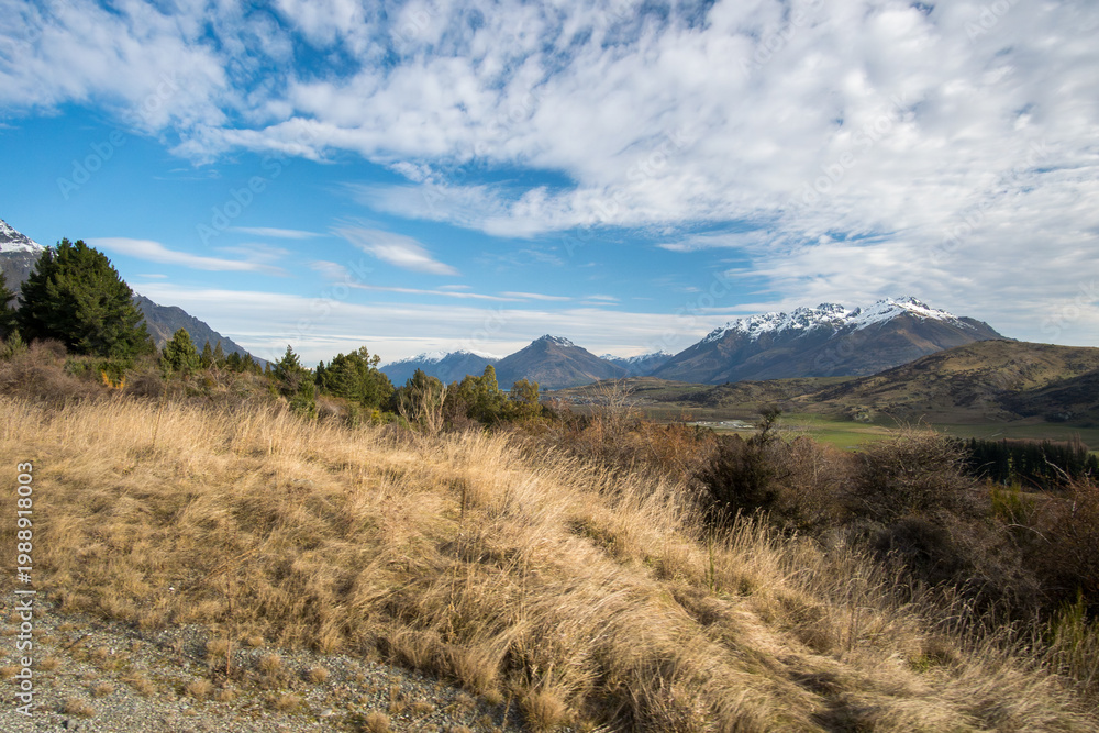 Fototapeta premium Paysage de prairie dorée aride avec montagnes enneigées et ciel bleu nuageux, région de Wanaka, île du Sud, Nouvelle-Zélande, automne