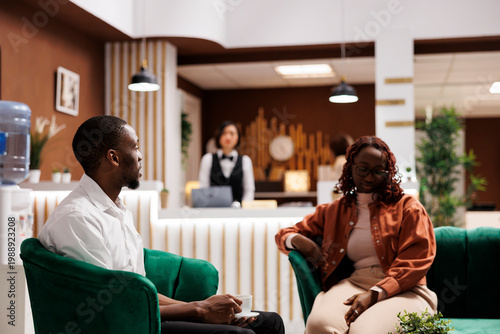 African american guests in lounge area talking and enjoying luxury service at hotel, waiting to fill in registration form for room reservation. Tourists sitting on sofa at front desk.