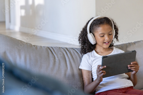 Fotografía African American female child sitting on sofa at home wearing headphones holding