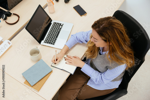 Woman writing in notebook while working with laptop
