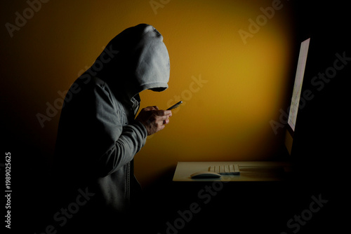 Foto Anonymous hacker using smartphone while working on computer in dark environment, representing mobile hacking, cybercrime and data theft
