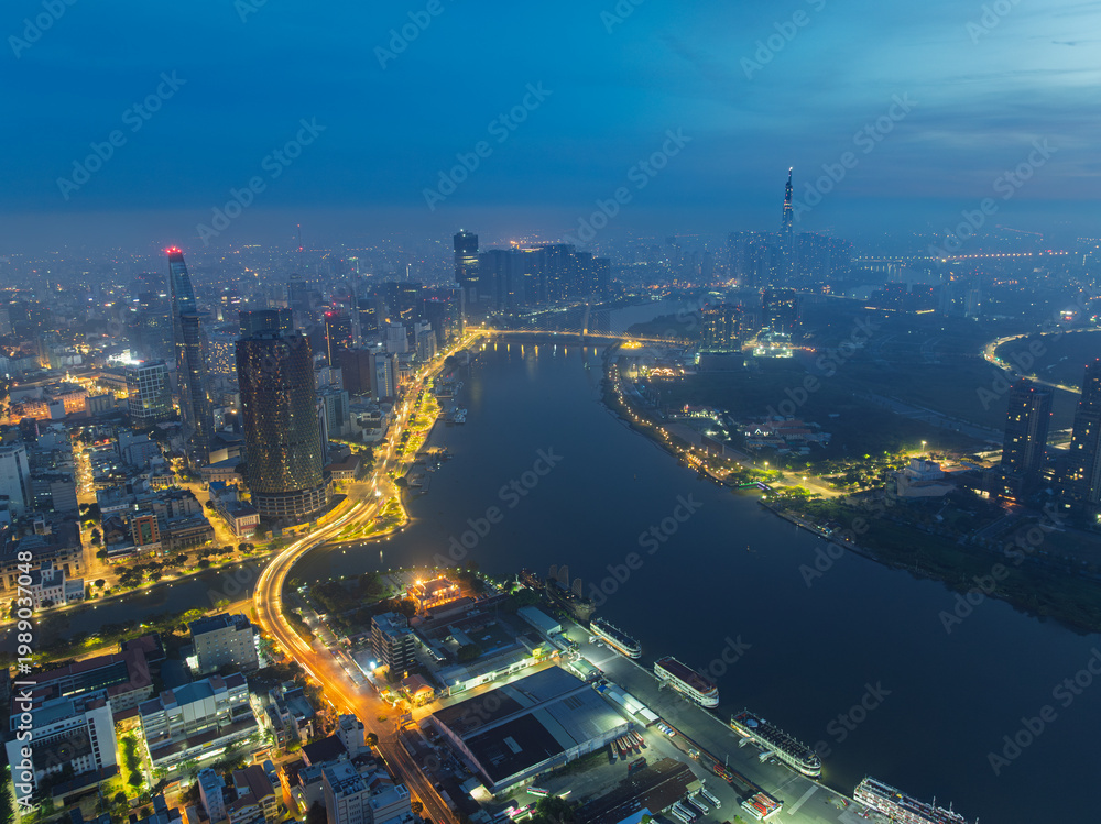 Obraz premium Aerial evening view over Saigon River with glowing roads, dense high-rise buildings, and atmospheric haze covering Ho Chi Minh City skyline.