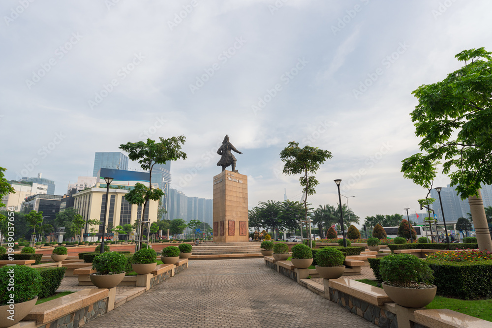 Obraz premium Tran Hung Dao statue monument centered in landscaped park with symmetrical pathways, trimmed shrubs, and modern skyline backdrop under cloudy sky in Ho Chi Minh City.