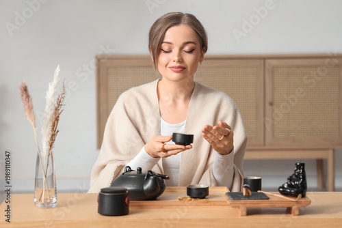 Beautiful young woman with cup of aromatic tea sitting at table in light room