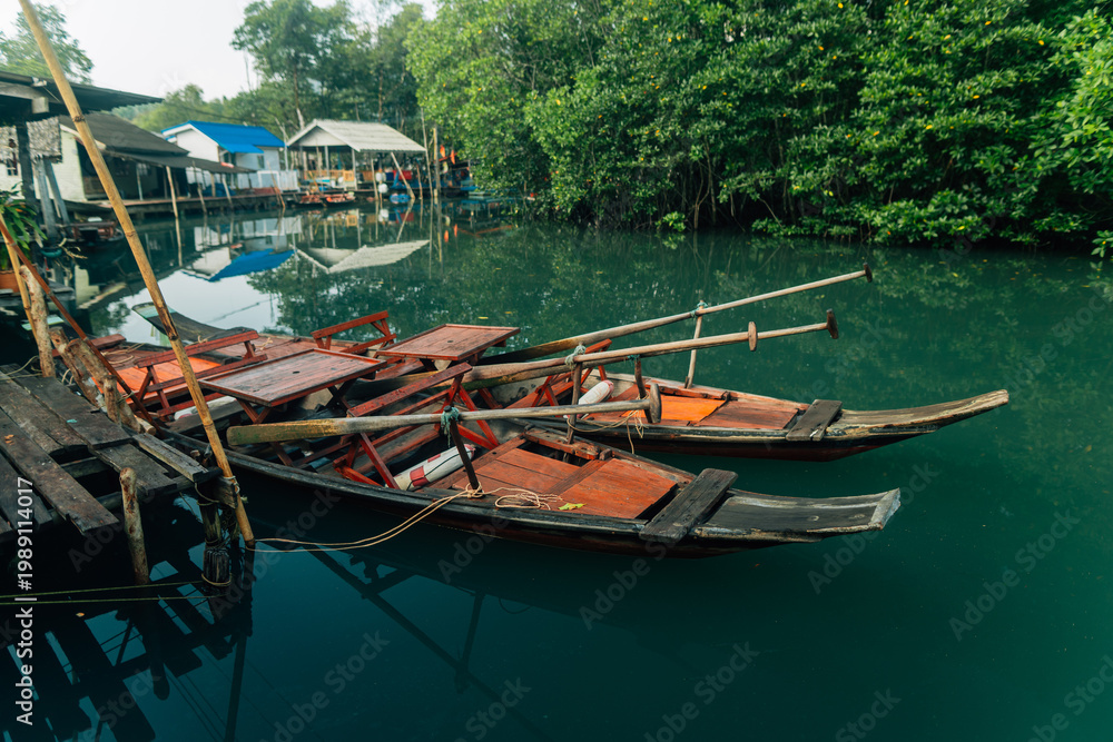 Naklejka premium Traditional wooden boats and stilt houses along a calm canal surrounded by lush green mangrove forest.