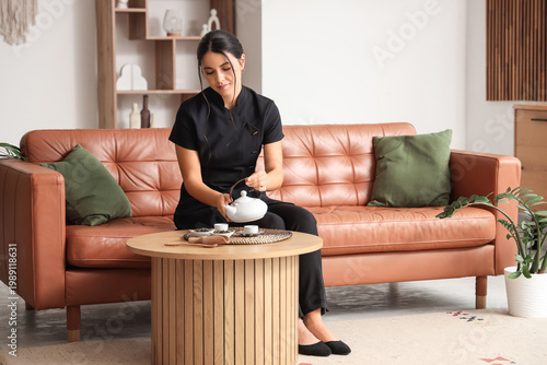 Beautiful young woman pouring tea from teapot at table in room