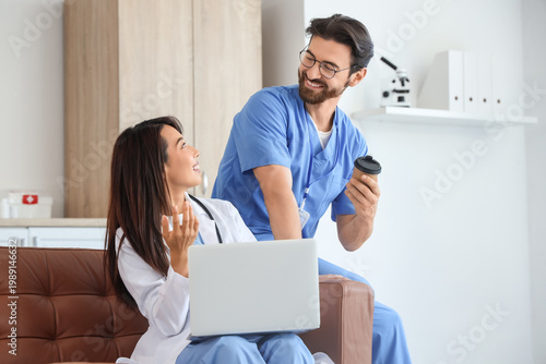Young doctors with coffee and laptop sitting on sofa in clinic