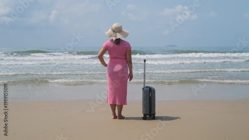 Back view of young woman standing near suitcase on sandy beach and watching ocean waves, enjoying long awaited vacation, slow motion. Travel relaxation concept