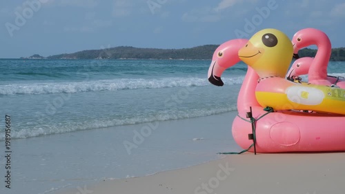 Pink inflatable flamingo and colorful rings placed on a wide sandy beach with rolling sea waves and tropical greenery in the background. Nature and leisure concept