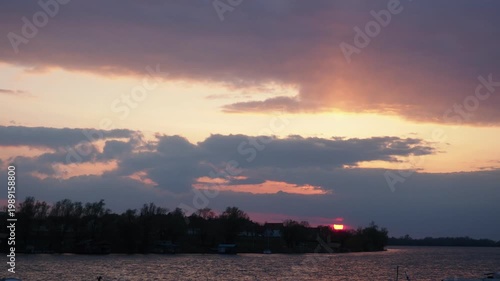 The sun sets over the lake as clouds fill the sky. Colors change from orange to purple. The view shows the lake shore and trees just before nightfall.