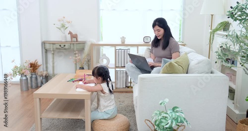 Asian single mother working on laptop from sofa while preschool daughter draws at coffee table, wide home learning scene with parent supervision, early education, childcare support, warm family bond