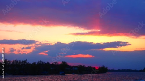 The sky changes colors as the sun sets over the water. Clouds float above while silhouettes of trees line the horizon. The scene shows a calm evening time by the lake.