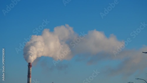 Smoke billows from a tall factory chimney as clear blue sky surrounds it. The activity takes place in an industrial area during late afternoon.