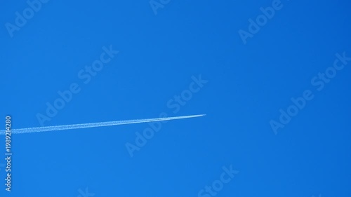 An airplane travels across clear blue sky leaving behind white trail. Plane moves swiftly against the backdrop of the city below. A perfect day for flying is shown here. Chemtrail conspiracy theory.