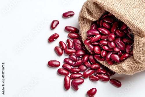 Red kidney beans spilling from a rustic burlap sack onto a white surface, overhead view of raw legumes.