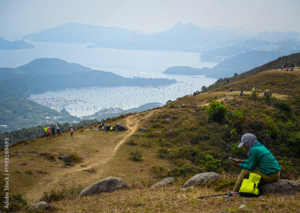 Fototapeta premium Hong Kong hikers resting on a hilltop overlooking a coastal bay with islands and boats in the distance.