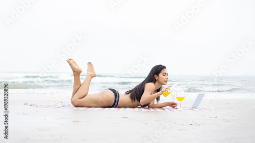 Obraz na plátně Southeast Asian woman relaxes on a sun-drenched beach, enjoying a chilled juice and creating a relaxed mood while wearing a bikini in this beachfront setting, featuring a blue ocean and sandy shore