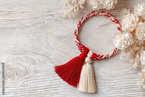 A handcrafted red and cream tassel ornament rests gracefully on a weathered wooden surface, complemented by a delicate arrangement of dried white flowers.