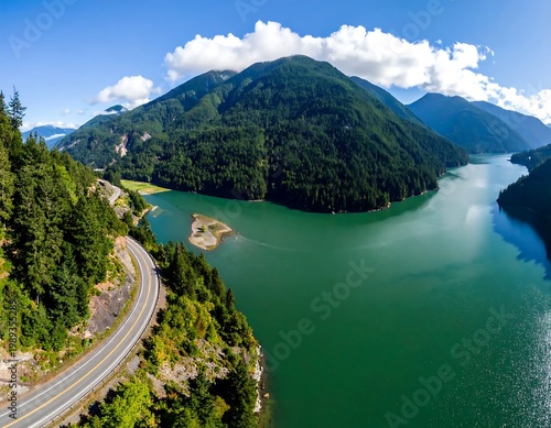Aerial Landscape Of A Winding Asphalt Road Alongside A Turquoise Mountain Lake Surrounded Dense Evergreen Forests Under A Sunny Blue